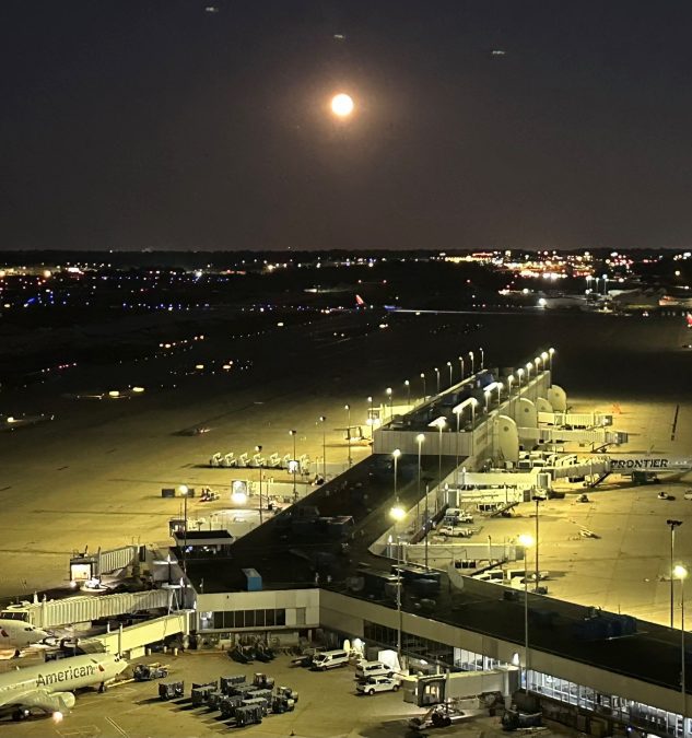 An aerial view of the airport terminal illuminated by a radiant super blue moon, casting a gentle glow over the scene. The moon dominates the night sky, appearing larger and brighter than usual, enhancing the contrast between the lit terminal structures and the surrounding darkness. Airplanes are parked at the gates, and runway lights twinkle in the distance, all under the mesmerizing presence of this rare lunar event.