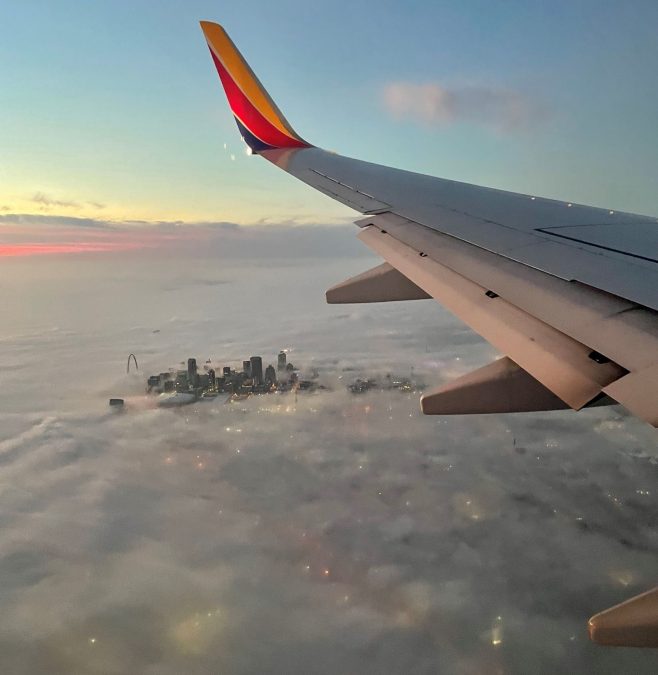 An aerial view shows a stunning sunrise over St. Louis, with the iconic Gateway Arch and downtown skyline peeking through a thick blanket of clouds. The scene is framed by the wing of a Southwest Airlines plane, highlighting the serene beauty of the moment as city lights glimmer faintly beneath the clouds. The colorful horizon signals the start of a peaceful journey.