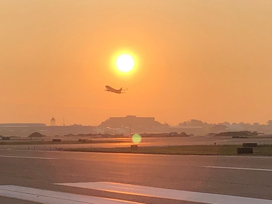 A plane ascends gracefully into the sky as the sun rises in the background, casting a warm golden glow across the runway at St. Louis Lambert International Airport. The early morning haze softens the light, creating a serene and peaceful atmosphere. The scene embodies the beginning of a new day filled with travels and opportunities.