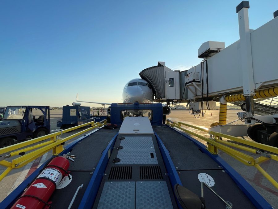 A ground-level view of a passenger plane parked at a gate, connected to a jet bridge under a clear blue sky. Yellow and blue ground equipment frames the scene, with a loading ramp leading directly toward the aircraft's front. The scene captures the preparation process before boarding or cargo handling at the terminal.