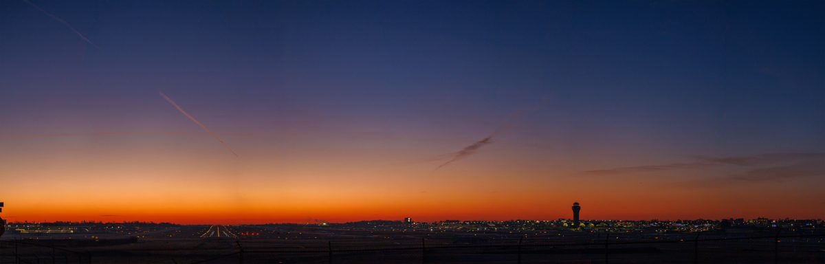 A panoramic view of St. Louis Lambert International Airport at dawn. The sky transitions from deep blue at the top to vibrant orange near the horizon, highlighting the airport's control tower and runway lights. The calm, early morning atmosphere showcases the city's skyline and airport operations waking to a new day.