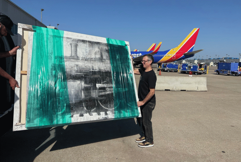 New artwork by Chris Day being delivered onto the grounds by two men, with planes in the background.