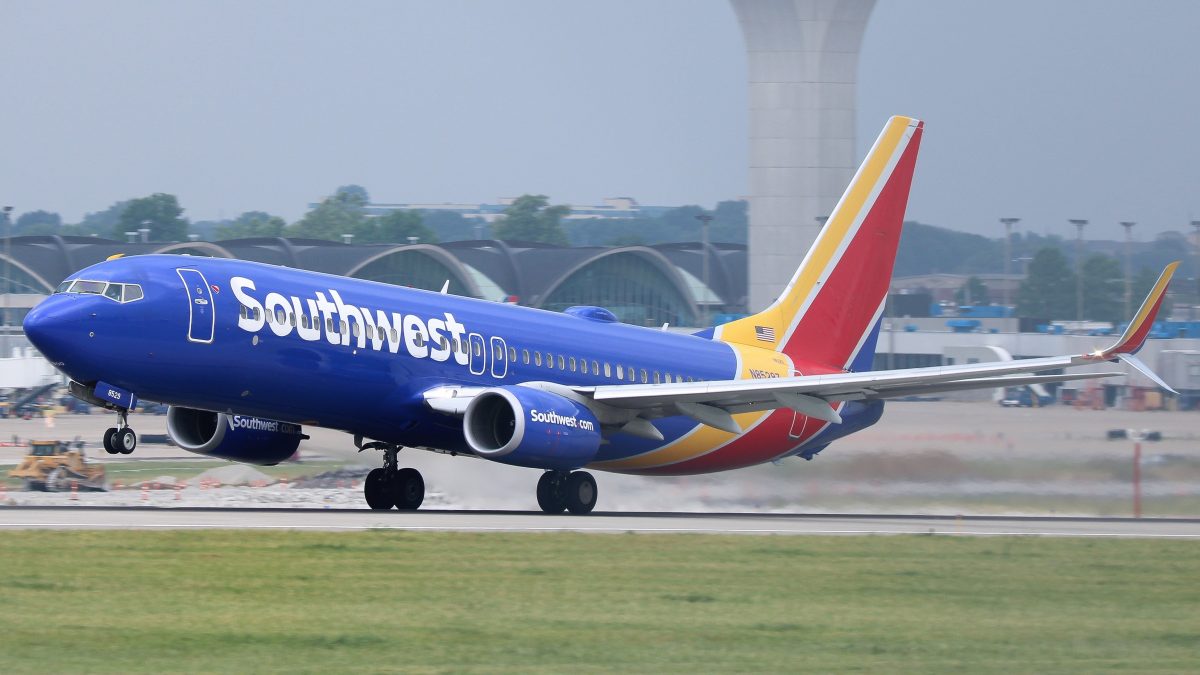 A Southwest Airlines Boeing 737 touches down on the runway, with its wheels just beginning to make contact. In the background, the iconic curved roof terminals and the air traffic control tower at St. Louis Lambert International Airport are visible. The plane's vibrant blue fuselage with red and yellow accents adds dynamic contrast against the grey skies and runway landscape.