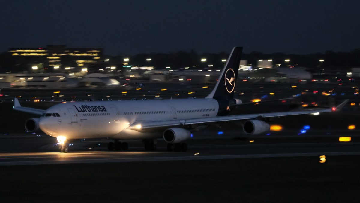 A Lufthansa long-range, wide-body aircraft prepares for departure at night, with its lights shining brightly on the runway. Surrounding airport lights and distant terminal structures create a sparkling backdrop against the dark sky. The sleek jet is poised for takeoff, emphasizing the readiness for international travel.