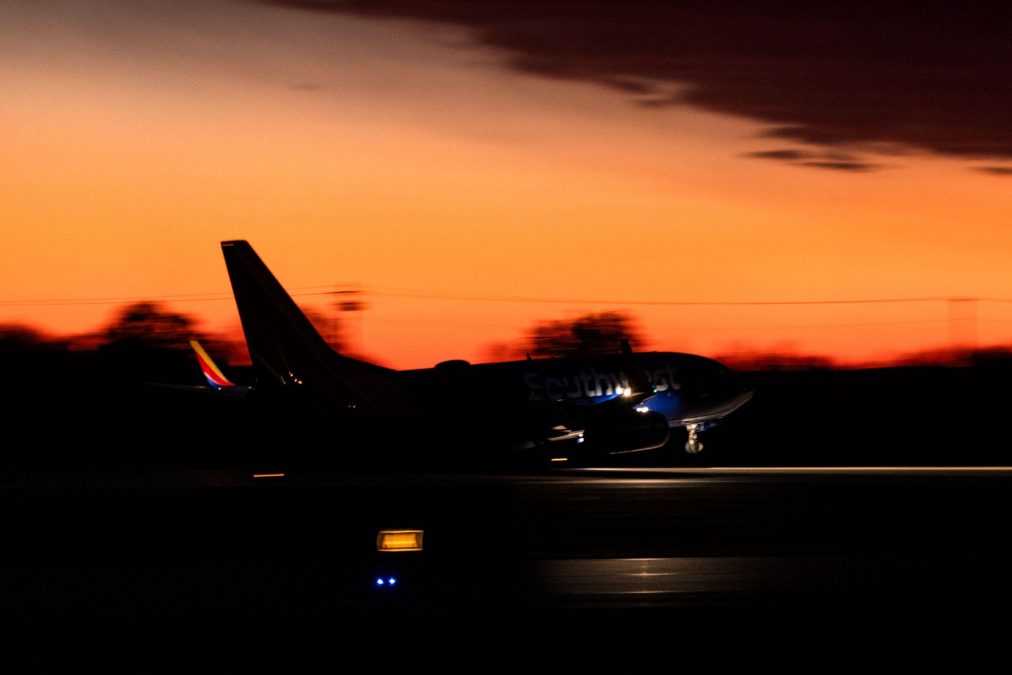 A Southwest Airlines plane speeds down the runway in silhouette against a stunning sunset sky. The deep orange and red hues blend into the darkening horizon, creating a dramatic contrast as the aircraft's colors and branding are subtly illuminated. The runway lights provide additional visual accents to the scene.