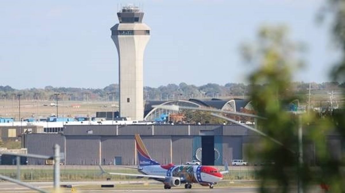 A Southwest Airlines plane featuring a decorative livery is positioned on the runway in front of St. Louis Lambert International Airport's signature air traffic control tower. The airport's arched terminal building is visible behind the tower, with a touch of greenery in the foreground framing the scene.