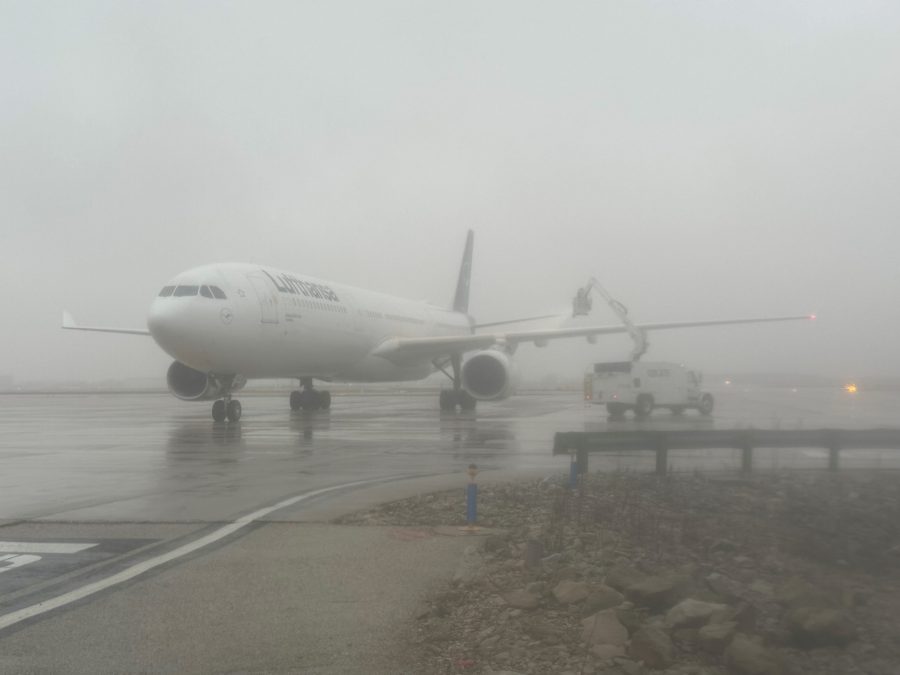 A Lufthansa Airbus A330 aircraft sits on a wet tarmac at St. Louis Lambert International Airport, surrounded by dense fog. The aircraft's nose is pointed slightly to the left, with its extended wings stretching into the misty background. A deicing truck with an elevated boom and a worker inside the enclosed platform is positioned near the right wing, spraying deicing fluid. The reflective surface of the tarmac and low visibility conditions emphasize the foggy atmosphere. Faint airport lights in the distance create a subtle glow, adding to the scene's moody, overcast feel.