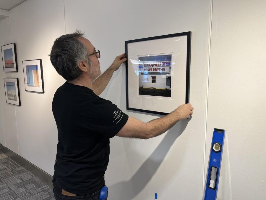 A man with short gray hair, glasses, and a black t-shirt is hanging a framed photograph on a white gallery wall. The photograph within the frame features a sign that reads 'DONUT DRIVE-IN' in bold letters. The man carefully adjusts the frame to ensure it is level. A blue leveling tool leans against the wall nearby. Other framed photographs are visible on the adjacent wall, showcasing a minimalist architectural theme. The setting is a modern exhibition space with a gray carpeted floor, white walls, and bright lighting, indicating an art installation in progress.
