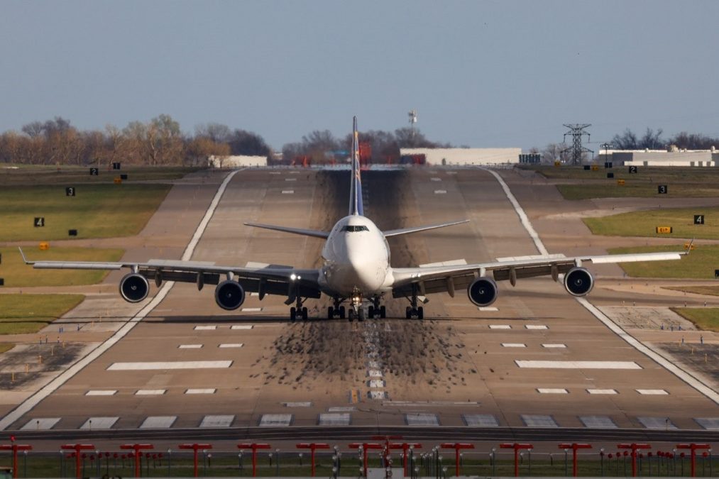 A large commercial aircraft is positioned on a runway at St. Louis Lambert International Airport, captured head-on. The plane's landing gear is fully extended, and the wide wingspan and engines are clearly visible. The runway stretches into the distance, with runway markings, lighting systems, and surrounding grassy areas creating a symmetrical view.