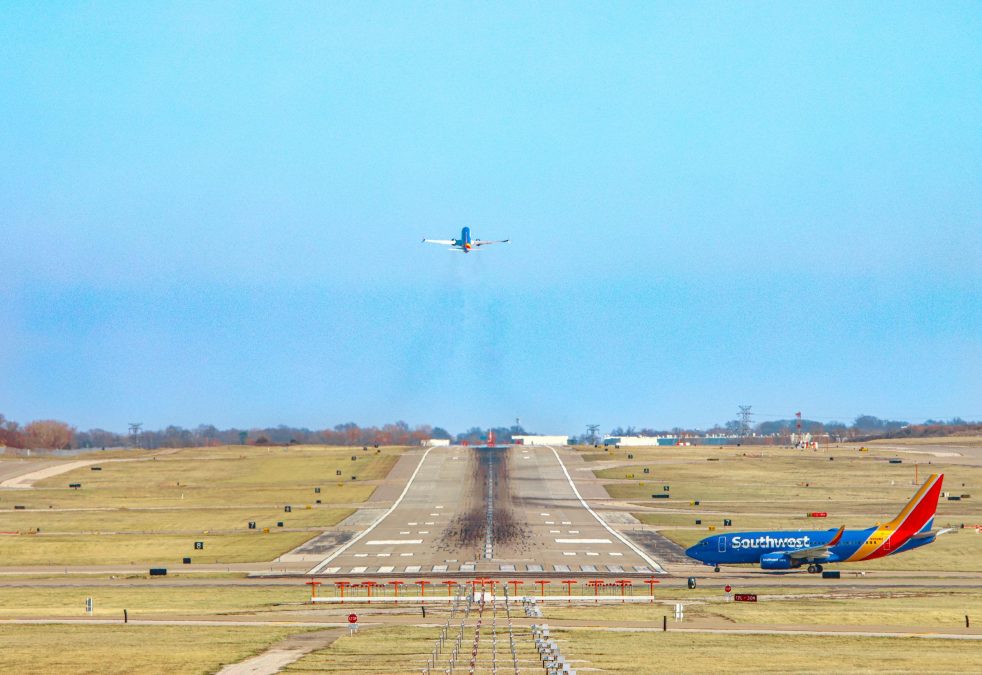 A wide view of a runway at St. Louis Lambert International Airport shows a Southwest Airlines plane taking off into a clear blue sky in the distance. In the foreground, another Southwest aircraft waits on a taxiway. The scene captures the vast open airfield with grassy areas on both sides of the runway and runway lighting equipment positioned near the camera.