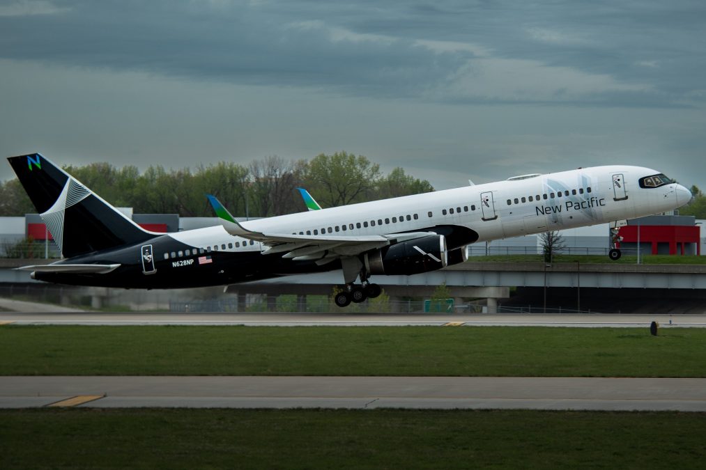 A New Pacific Airlines plane is taking off from St. Louis Lambert International Airport. The aircraft, with a sleek white and black color scheme and the airline's name displayed on the fuselage, is captured mid-lift, with its landing gear extended. The overcast sky and surrounding greenery create a dynamic backdrop for the scene.