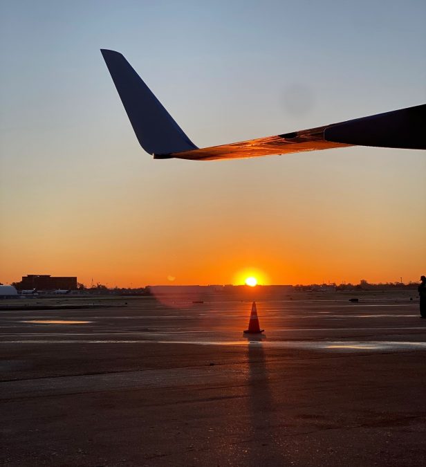 A stunning sunrise at St. Louis Lambert International Airport is captured with the silhouette of an aircraft's wing in the foreground. The sun is low on the horizon, casting a warm, orange glow across the tarmac. A traffic cone positioned below the wing creates a long, distinct shadow stretching toward the viewer.