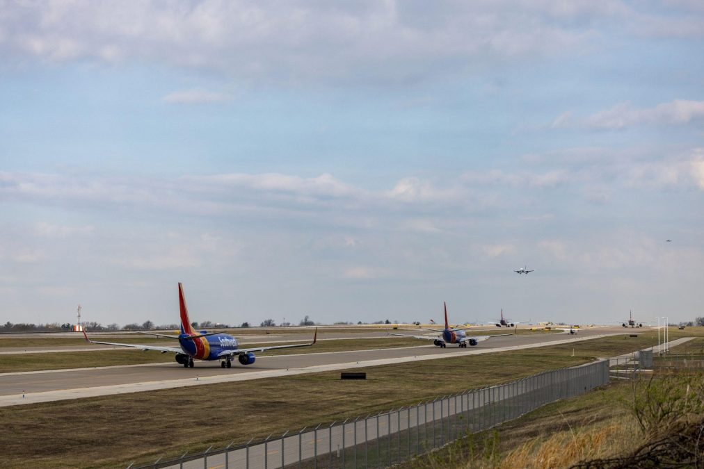Several Southwest Airlines planes line up on taxiways at St. Louis Lambert International Airport, awaiting departure. In the distance, a plane is on approach for landing under a partly cloudy sky. The scene shows the orderly flow of air traffic with the airfield's grassy areas and fencing visible in the foreground.