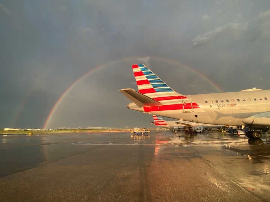 A vibrant rainbow stretches across the sky at St. Louis Lambert International Airport, with a secondary, faint rainbow visible alongside it. In the foreground, multiple American Airlines planes are parked on the wet tarmac, their reflective surfaces catching the warm light after a rainstorm. The colorful scene contrasts against the moody, dark clouds in the background.