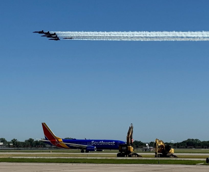 A formation of military jets performs a flyover at St. Louis Lambert International Airport, leaving white vapor trails across the clear blue sky. Below, a Southwest Airlines plane is parked on the tarmac near two pieces of construction equipment.