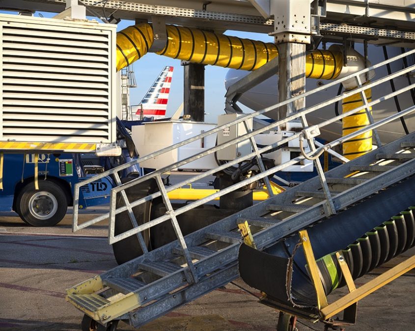 A detailed view of airport ground operations with equipment surrounding a jet on the tarmac. A bright yellow ventilation hose curves across the scene, while a metal conveyor belt is positioned for cargo loading. In the background, part of an American Airlines plane's vertical stabilizer is visible, adding a subtle yet recognizable element.