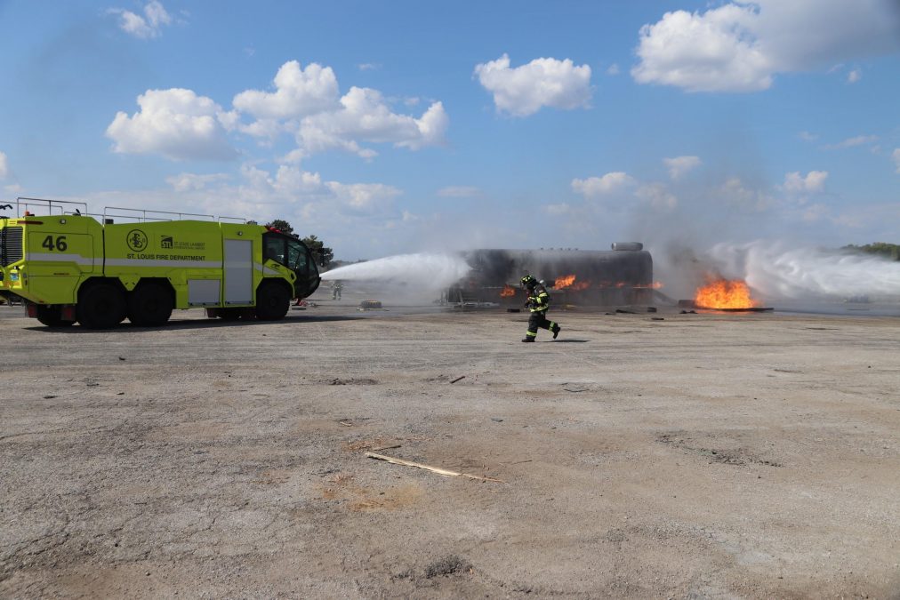 A bright yellow St. Louis Lambert Airport Fire Department truck, marked with the number 46, sprays powerful jets of water toward a large burning structure in an outdoor training exercise. Flames and smoke billow across the scene under a clear blue sky, while a firefighter in full gear runs across the foreground, emphasizing the intensity of the drill.