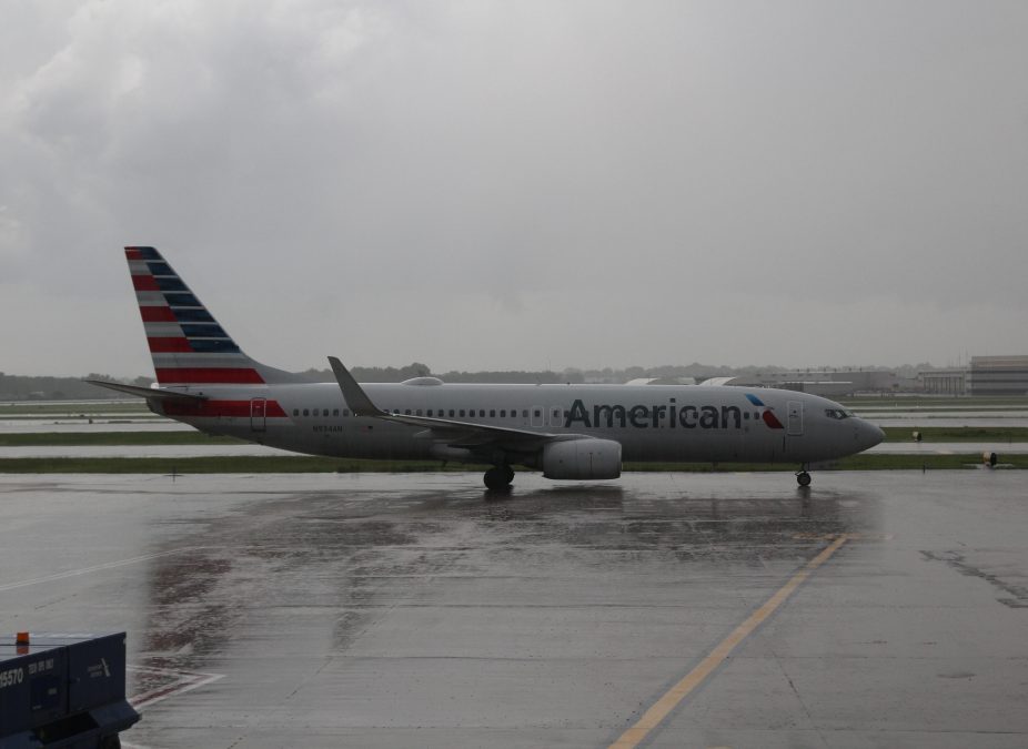 An American Airlines plane is parked on a wet tarmac during a rainy, overcast day. The aircraft's iconic red, white, and blue tail design stands out against the gray sky. Reflections from the rainwater create a glossy surface on the ground, highlighting the somber atmosphere of the scene.
