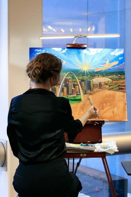 An artist paints with her back to the camera, a stunning portrait of downtown St. Louis.