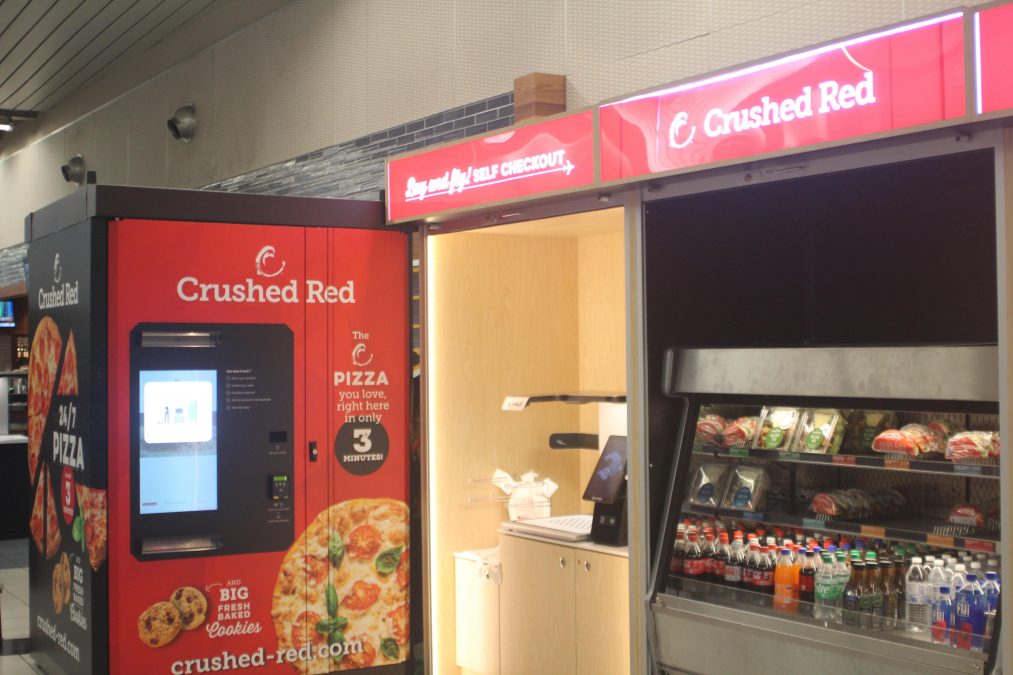 A Crushed Red self-service kiosk at St. Louis Lambert International Airport offers quick meals, including pizzas that can be ready in three minutes. The kiosk features bright red branding and signage. Refrigerated shelves display prepackaged salads, sandwiches, and beverages, with a self-checkout station available for easy purchases.