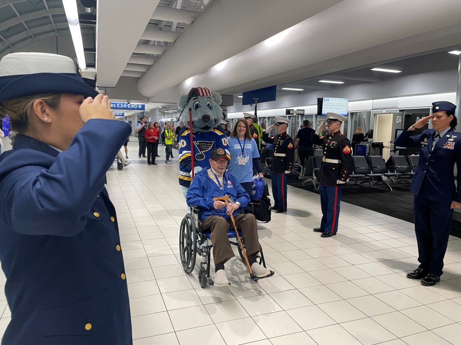 Blues Mascot Bluey wheels a veteran in. wheelchair through the airport while airmen and women salute.