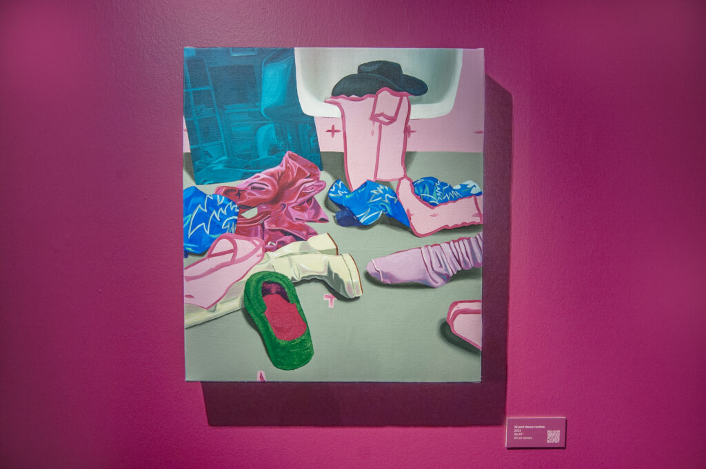This image shows a vibrant painting mounted on a magenta wall, titled "32 part dance routine." The artwork features a playful arrangement of clothing items, including colorful socks, a cowboy boot, and scattered garments, with a black hat resting on a chair in the background. The vivid colors and abstract composition give the piece a dynamic, expressive feel, inviting the viewer to imagine the story or movement behind the scene. A plaque with the artwork's title and a QR code is positioned to the right.