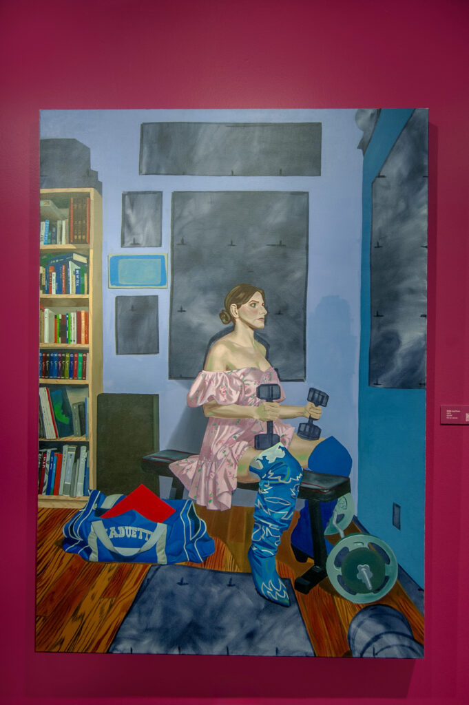 510 lb leg lift by Rachel Lebo. This painting depicts a woman seated on a workout bench, holding dumbbells, and wearing a pink floral dress with patterned blue boots. Surrounding her are exercise equipment, including a barbell plate and a duffel bag. Bookshelves filled with various books line the left side, while a series of framed gray panels adorn the background.
