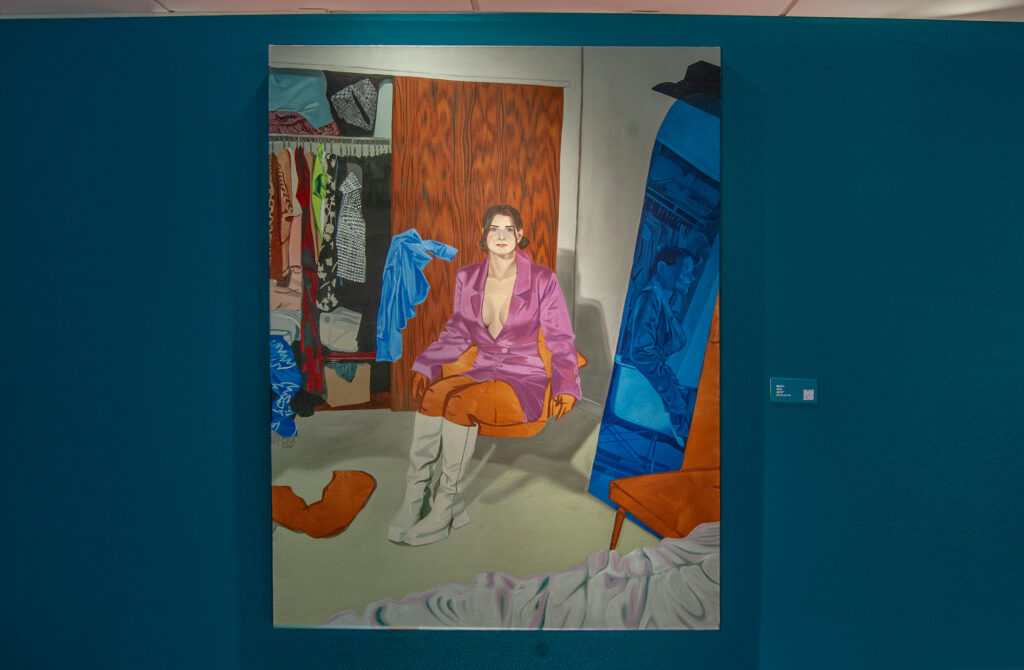 This painting, titled "In appreciation of Pam, Kim, and Dolly," is displayed on a teal wall. It depicts a woman sitting on an orange chair in a closet or dressing room, wearing a vibrant pink jacket and white knee-high boots. Behind her, a mirror reflects a monochromatic blue-toned version of her image. To the left, colorful clothing hangs in an open wardrobe, adding a dynamic mix of patterns and textures.