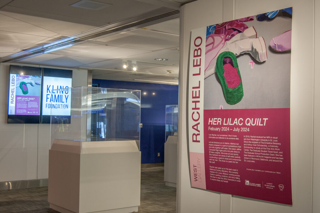This image highlights an exhibition space promoting "Her Lilac Quilt" by Rachel Lebo, on display from February to July 2024. A large, colorful poster showcases artwork from the series, featuring a green slipper and scattered clothing items. The accompanying text provides background on Rachel Lebo, her artistic inspiration, and her education, noting her MFA from Washington University in St. Louis. Digital displays and exhibit cases in the background further emphasize the gallery’s dedication to showcasing contemporary art and storytelling. The exhibit is sponsored by the Kling Family Foundation and part of the Lambert Art and Culture Program.