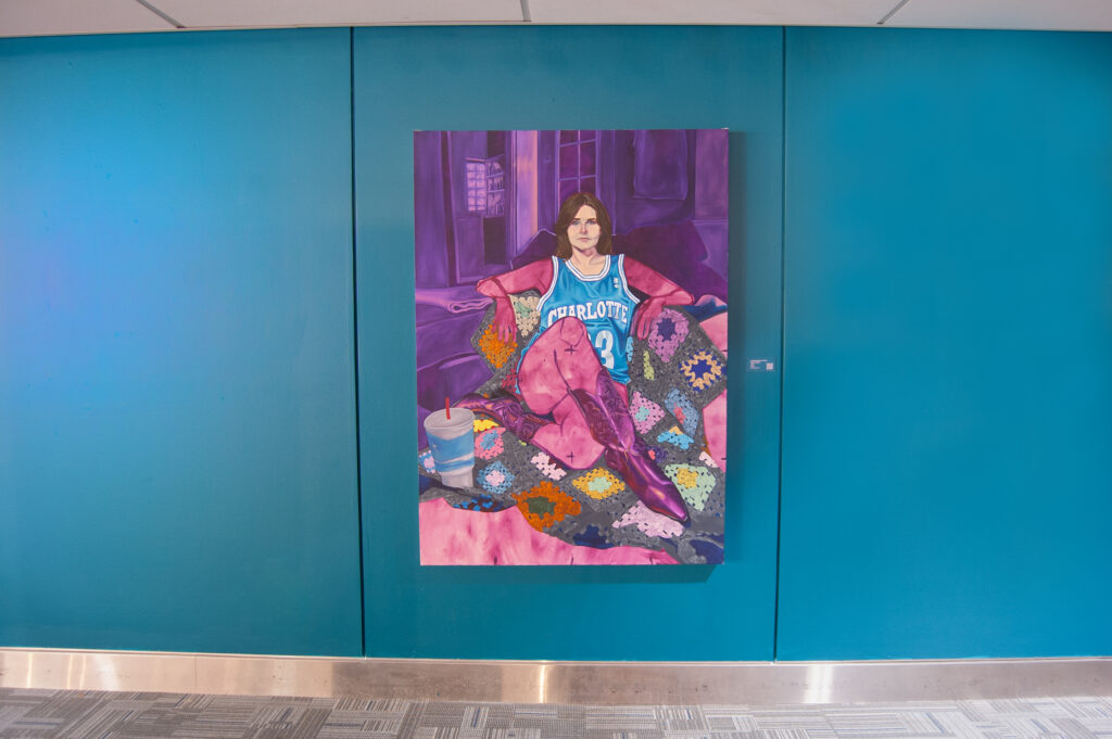 This image features a vibrant portrait painting mounted on a teal wall. The artwork depicts a person lounging on a colorful crochet blanket, wearing a Charlotte Hornets basketball jersey and purple boots. A drink cup with a straw rests nearby, adding a casual, relaxed atmosphere to the scene. The rich use of purples, blues, and other bold hues creates a lively and visually striking composition. The piece is "Marina" by Rachel Lebo