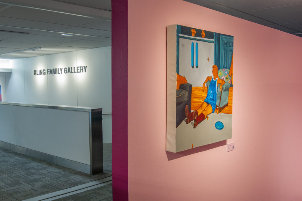 This image shows a painting, "Sitting in his chair" displayed on a light pink wall in the Kling Family Gallery. The artwork depicts a figure relaxing in an interior space, wearing a blue sports jersey and cowboy boots.