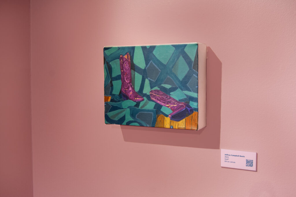 This image showcases a painting titled "Jeffrey Campbell Boots," displayed on a pink wall. The artwork features a pair of vibrant purple boots resting on a richly patterned teal and green surface. The bold contrast between the boots and the abstract background emphasizes the playful and stylish composition. A plaque with the title, date (2023), medium (oil on canvas), and a QR code is positioned to the right of the painting, inviting viewers to explore more about the piece.