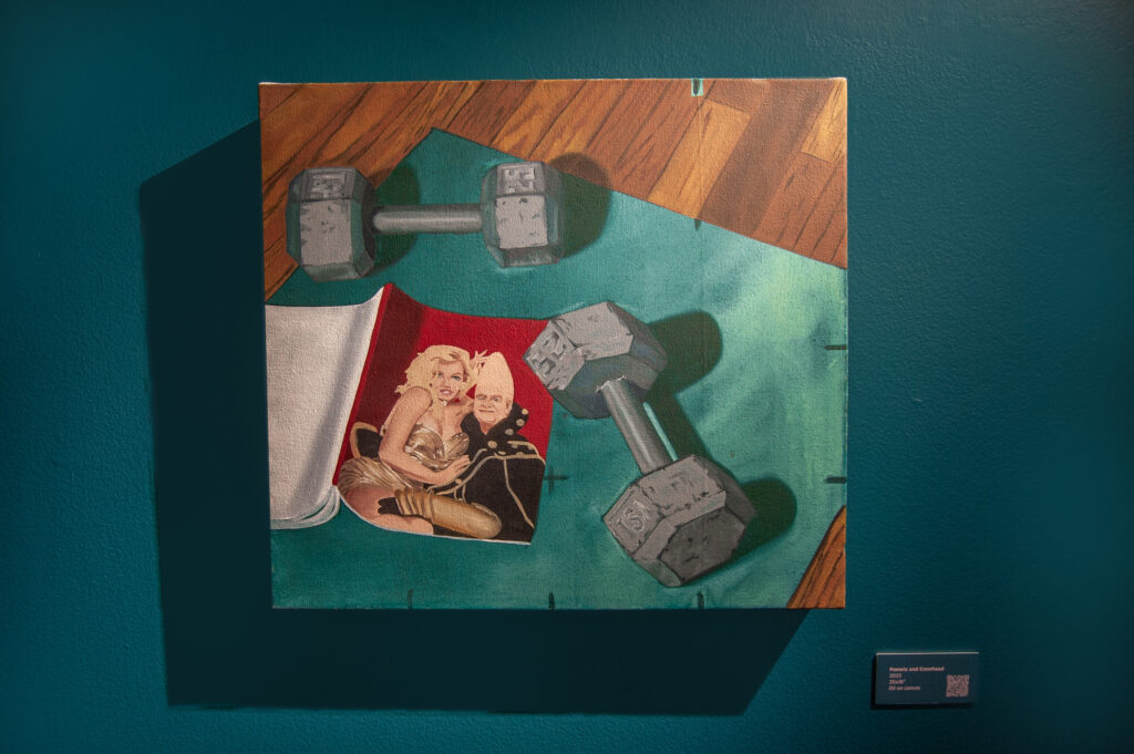 This painting, titled "Pamela and Conehead," is displayed on a teal wall. The artwork features a scene with two dumbbells resting on a green surface, alongside a magazine showing Pamela Anderson embracing a character from the "Coneheads" movie.
