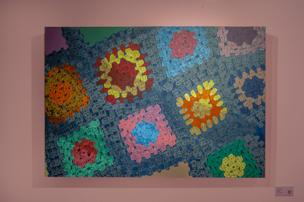This painting highlights a large, colorful crochet-inspired design, displayed on a pale pink wall. The textured blue background enhances the depth and complexity of the piece, capturing the warmth and charm of traditional crochet craftsmanship. A small plaque with a QR code is positioned on the right for further details.