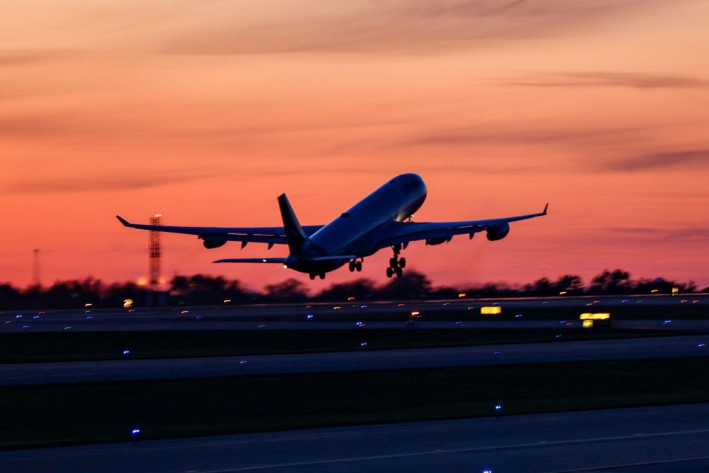 An aircraft is captured mid-takeoff at St. Louis Lambert International Airport, silhouetted against a vibrant orange and pink sunset sky. The runway lights glow in deep blue hues along the taxiway, contrasting with the fading light of dusk. Trees and distant airport structures form a dark horizon beneath the colorful sky.
