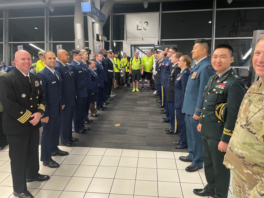 A line of uniformed military personnel stands at attention at St. Louis Lambert International Airport near Gate E8, forming a welcoming path. In the background, a group of Honor Flight volunteers, dressed in bright yellow shirts, prepares to escort veterans. The scene highlights respect and recognition for those who have served, with a mix of smiles and solemnity.