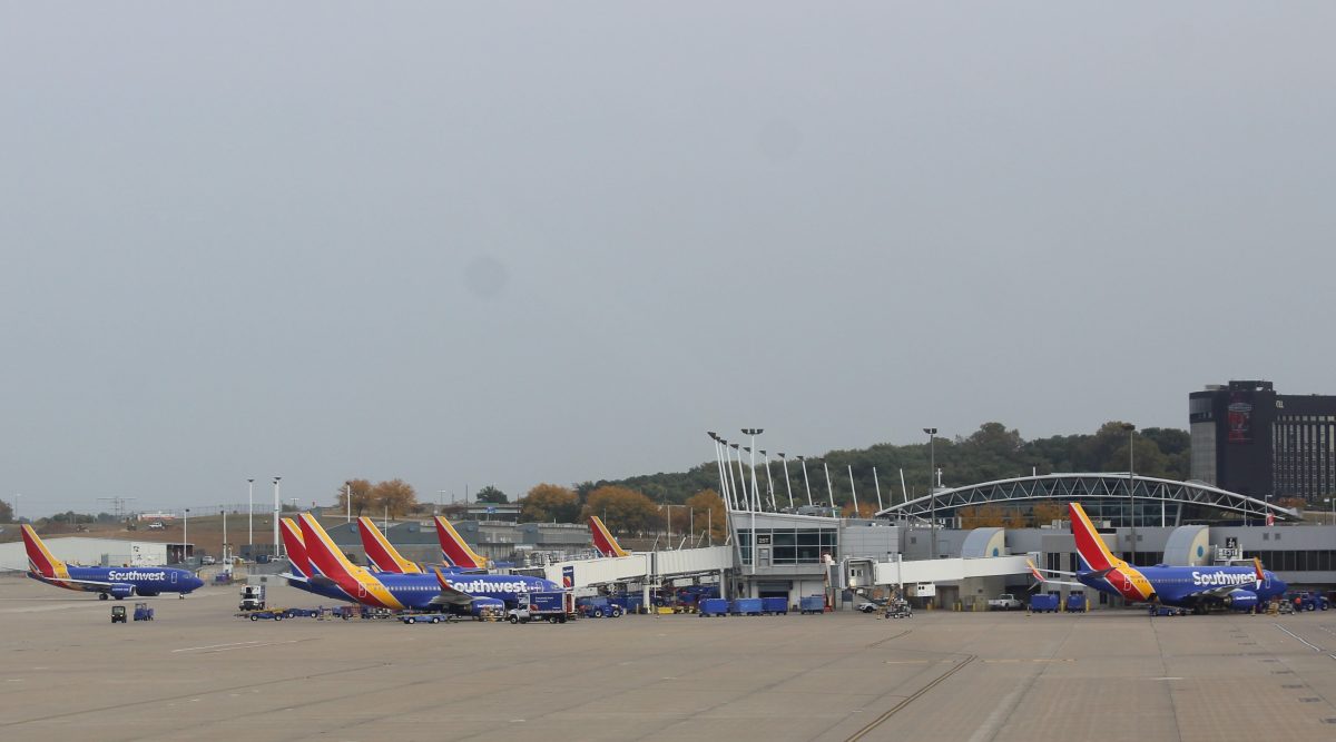 A row of Southwest Airlines planes is parked at the gates of an airport terminal, with their vibrant red, blue, and yellow tail fins creating a striking visual pattern. The terminal bridge and infrastructure are bustling with activity, including ground crew and equipment in motion. Trees with fall-colored leaves frame the background, while a tall building is visible in the distance.