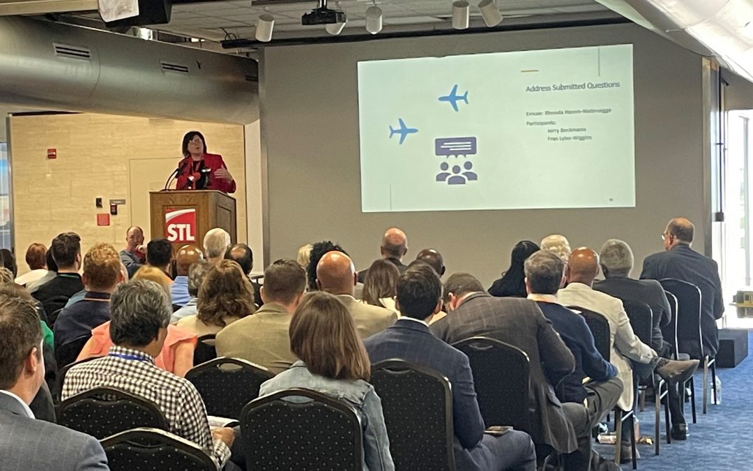 A speaker at the STL Industry Day event stands at a podium with the STL logo, addressing the audience. The presentation slide behind them has the heading "Address Submitted Questions" and includes airplane graphics and an icon representing a discussion. The audience is seated attentively, engaged in the session focused on airport terminal program updates.