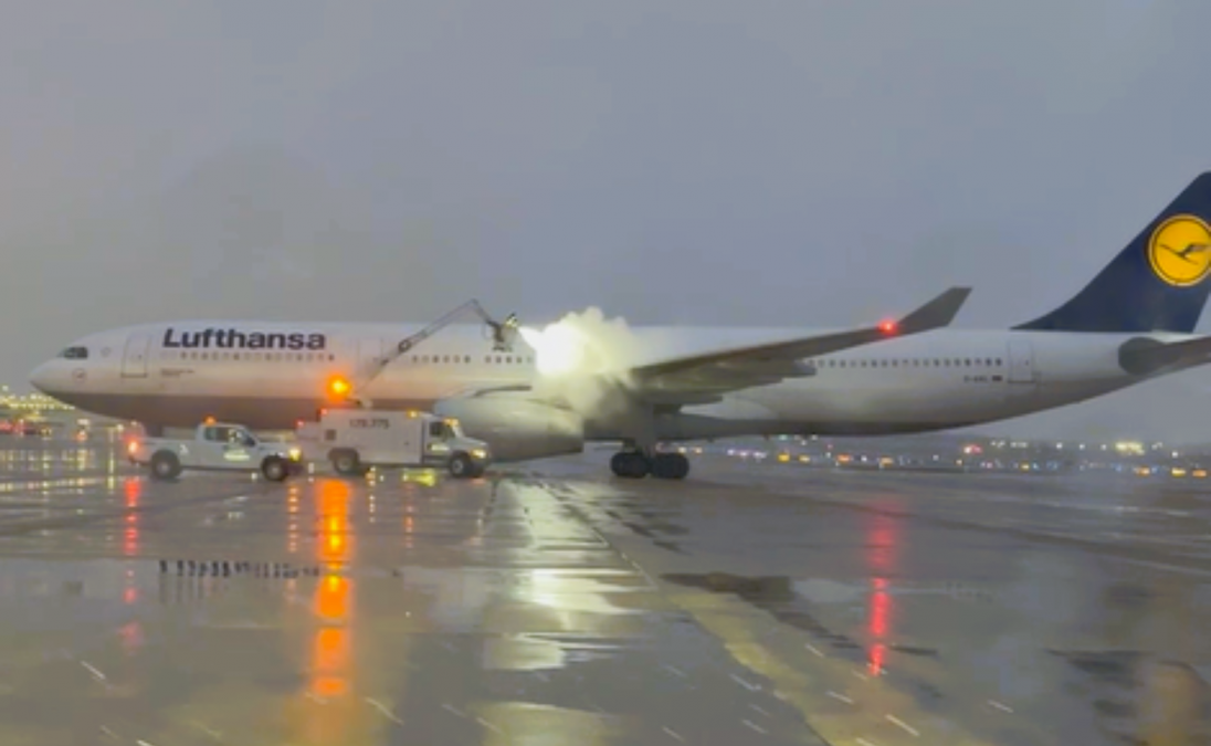 A Lufthansa aircraft undergoes deicing at St. Louis Lambert International Airport on a rainy, overcast day. Ground crew vehicles spray a heated deicing fluid onto the plane's wings and fuselage. Reflections from the airport lights glimmer on the wet tarmac, adding brightness to the otherwise dreary atmosphere.