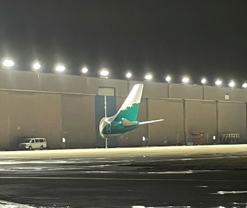 A large aircraft tail, painted green with white detailing, protrudes from a hangar at night. Bright overhead lights illuminate the scene, casting reflections on the wet ground. A white van parked nearby gives a sense of scale to the towering tail section.