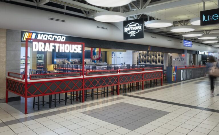 A dining area inside St. Louis Lambert International Airport features the NASCAR Drafthouse, marked by a large illuminated sign. The bar area includes high-backed chairs and a red railing with a crisscross design, with tire-themed decor and checkered flags enhancing the NASCAR theme. Overhead circular lights and nearby directional signage complete the modern airport setting.