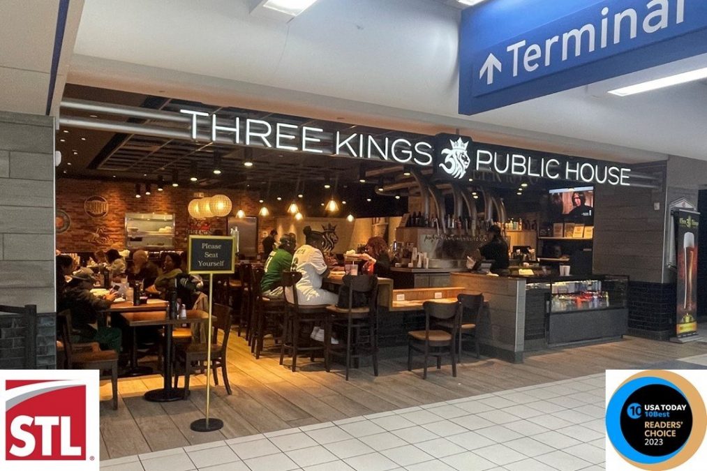 The entrance of Three Kings Public House at St. Louis Lambert International Airport showcases warm, inviting lighting with diners seated inside at both tables and the bar. The establishment's illuminated sign is displayed prominently above the entrance, and a "Please Seat Yourself" sign stands at the front. On the right side, a USA Today "10 Best Readers' Choice 2023" badge highlights its recognition.