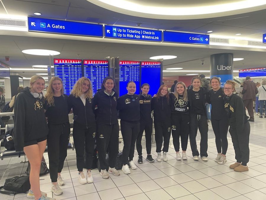 A group of athletes from the University of Colorado women's hockey team stands together at an airport terminal, posing in front of a large digital arrivals board. They are dressed in matching black team outfits with the "CU" logo prominently displayed. Overhead, directional signage points travelers toward ticketing, gates, and rideshare services.