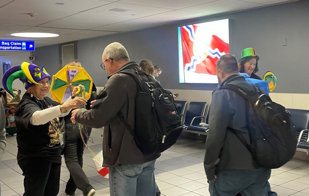 A lively Mardi Gras celebration takes place inside St. Louis Lambert International Airport as arriving passengers are greeted by volunteers dressed in festive attire. A woman wearing a purple, green, and yellow jester hat hands out colorful bead necklaces to a traveler carrying a backpack. Another volunteer, wearing a green glittery hat, smiles while holding additional decorations. Behind them, a large illuminated sign displays the flag of St. Louis. The setting includes airport seating and signage directing passengers to baggage claim and transportation, creating a welcoming and festive atmosphere for travelers.