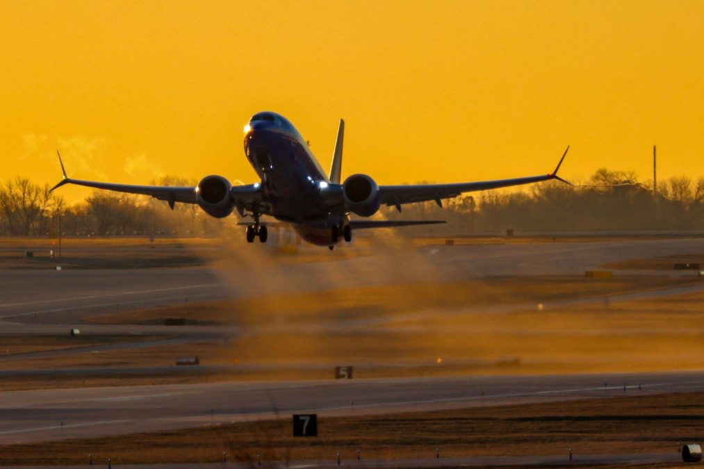 A commercial airplane takes off from St. Louis Lambert International Airport at sunrise, silhouetted against a golden sky. The aircraft, a Southwest Airlines Boeing 737, lifts off the runway with its landing gear still extended, trailing mist and vapor from the cold morning air.