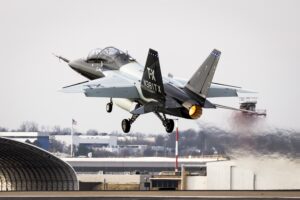 "High-resolution photograph of a Boeing T-X advanced trainer jet taking off from a runway with its afterburner engaged. The aircraft, designed for the U.S. Air Force, features a sleek, black-and-gray livery with 'TX' and 'N381TX' marked on its twin vertical stabilizers. The tandem-seat cockpit is occupied by two pilots wearing helmets and flight gear. Heat distortion from the engine is visible behind the aircraft as it ascends.