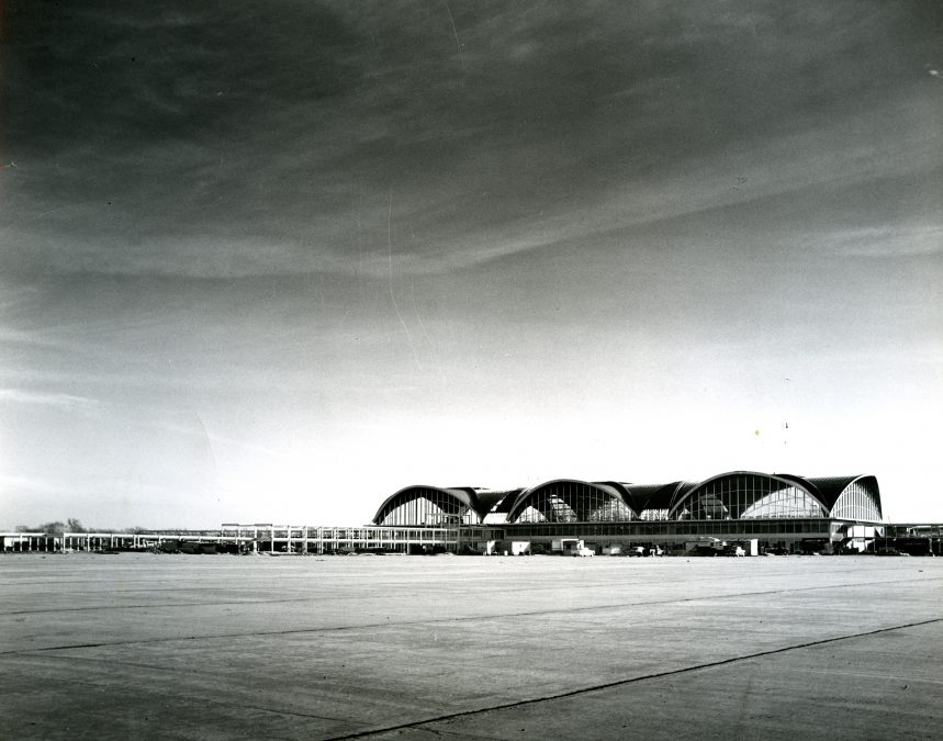 Black-and-white photograph of St. Louis Lambert International Airport’s iconic terminal, designed by architect Minoru Yamasaki. The image captures the terminal’s distinctive arched roof design, which features a series of sweeping, curved structures with large glass windows. Taken from a distance across the tarmac, the photograph showcases the expansive concrete apron in the foreground, with small vehicles and airport infrastructure visible near the terminal. The sky above is vast and open, adding to the grandeur of the architectural design.