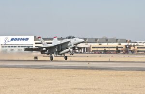 A U.S. Navy F/A-18F Super Hornet takes off from a runway outside the Boeing manufacturing facility in St. Louis. The aircraft, a twin-engine multirole fighter, is painted in a light gray camouflage scheme with red and black markings on the vertical stabilizers. Heat distortion from the jet engines is visible behind the aircraft as it lifts off the ground. The Boeing facility, with its large white sign displaying the company's blue logo, is prominently featured in the background. The surrounding area consists of dry grass, pavement, and parked vehicles, highlighting the active production and testing of military aircraft in St. Louis.