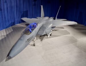 "High-resolution photograph of a Boeing F-15 Silent Eagle prototype displayed under bright lighting against a backdrop of blue curtains. The aircraft, an advanced variant of the F-15 fighter jet, features a sleek gray paint scheme with low-visibility markings. Its twin-seat cockpit is illuminated with blue lighting, showcasing the modern avionics and controls inside. The aircraft's conformal weapon bays and radar-absorbing coatings are part of its stealth enhancements.