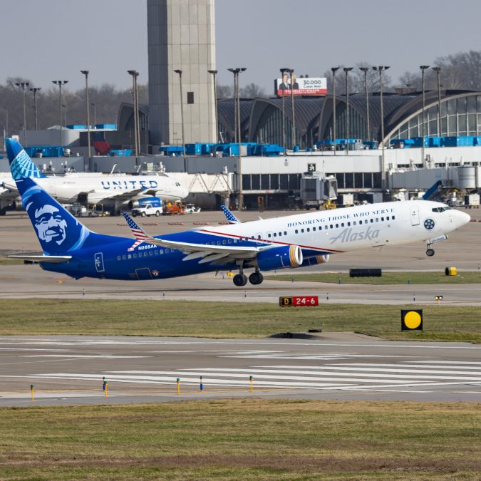 An Alaska Airlines jet painted in a special patriotic livery reading “Honoring Those Who Serve” is taking off from a runway. The aircraft features a bold blue tail with stars.